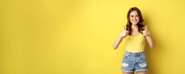 Portrait of stylish modern girl, feminine woman showing thumbs up, recommending gesture, like or approve, praise, standing over yellow background.