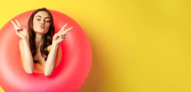Portrait of attractive modern girl inside trendy pink beach swimming ring, showing peace v-sign and smiling happy, standing over yellow background.