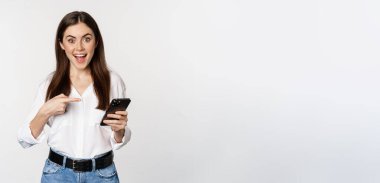 Enthusiastic girl pointing finger at smartphone and smiling, showing big sale online, using app on mobile phone, white background.