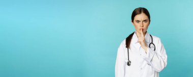 Angry female doctor, healthcare medical worker shushing with disapproval, taboo quiet gesture, silence someone, standing over torquoise background.