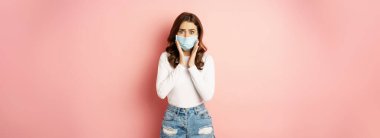Covid-19 and quarantine concept. Stylish girl in medical face mask, looking worried and shocked at camera, standing against pink background.