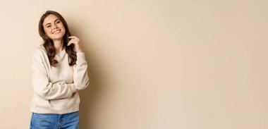 Beautiful young brunette woman posing happy against beige background, smiling at camera, wearing sweater.