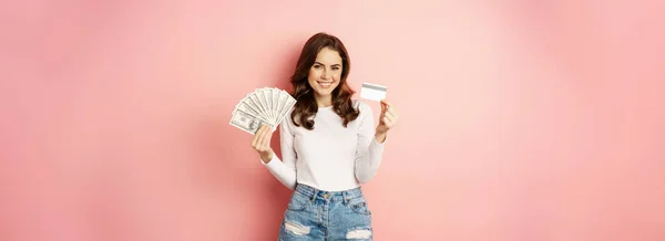 お金および貸付け金の概念 幸せな美しい女の子を保持クレジットカードと現金 満足そうな ショッピングを考えて ピンクの背景の上に立って — ストック写真
