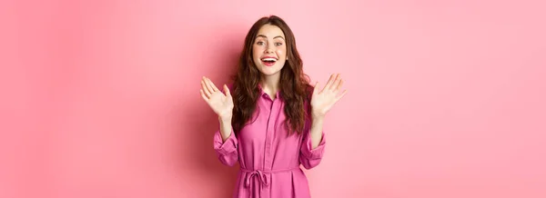 快乐的黑发女子的画像举手表决 接受礼物 得意洋洋地站在粉红的背景上 — 图库照片