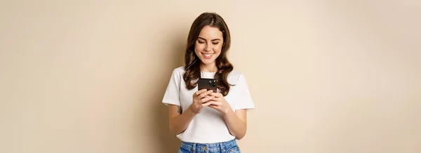 年轻的现代女孩在应用程序上聊天 使用智能手机应用程序和微笑 站在米色背景之上 — 图库照片