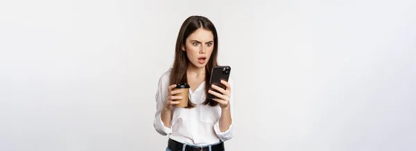 女性のコーヒーカップを保持し ショックを受けてスマートフォンを見て 唖然と顔 白い背景の上に立っている スペースのコピー — ストック写真