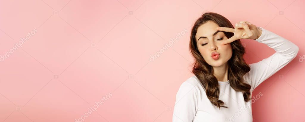 Close up portrait of coquettish young woman smiling, showing peace, v-sign gesture and posing happy, standing against pink background.
