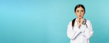Worried and concerned physician, hospital worker in white lab coat, looking scared and nervous at camera, standing with stethoscope over torquoise background.