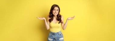 Woman shrugging shoulders and looking clueless, apologizing and smiling awkward, dont know, puzzled, has no idea, standing against yellow background.