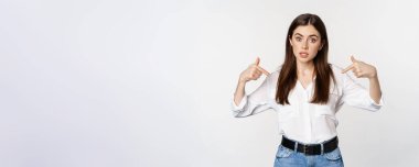 Portrait of young woman pointing at herself, self-promoting, standing over white background. Copy space