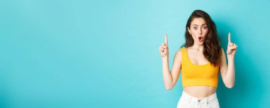 Yaz yaşam tarzı konsepti. Şaşırmış genç bir kadın heyecanla nefes nefese, parmaklarını havaya kaldırarak, hayrete düşmüş bir şekilde reklamını yapıyor, mavi arka planda duruyor..