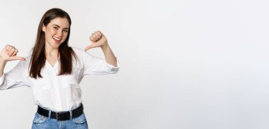Portrait of young woman pointing at herself, self-promoting, standing over white background. Copy space