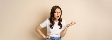 Confused woman looking clueless and annoyed, cant understand, asking whats problem, standing over beige background.