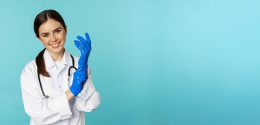 Smiling young woman, medical worker wearing gloves for patient checkup, standing in hospital clinic uniform over blue background.
