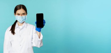 Angry and confused woman doctor in face mask, gloves, showing smartphone app, mobile screen, frowning upset, standing over blue background.