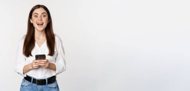 Young woman with smartphone, smiling and looking at camera, using mobile phone app, cellular technology and online shopping concept, white background.
