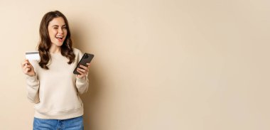 Smiling girl using mobile app, smartphone shopping and credit card, standing over beige background, order smth.