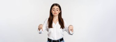 Disappointed young woman pointing fingers down, looking below with regret and sadness, standing in blouse and jeans over white background.