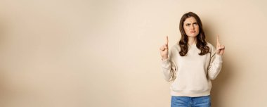 Confused young woman pointing fingers up and staring puzzled, cant understand promo text upwards, beige background.