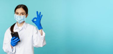 Online doctor and clinic. Young woman in medical face mask, using smartphone for client remote online appointment, showing okay sign, standing over blue background.
