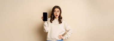 Woman showing smartphone screen and looking surprised at mobile phone, standing in sweater over beige background.