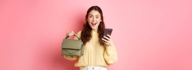 Heyecanlı esmer kız, elinde akıllı telefonuyla online mağazadan çanta sipariş etti. Yeni çantasını gösteriyor ve şaşkın bir şekilde gülümsüyor. Pembe arka planda duruyordu..