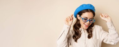 Portrait of stylish happy woman dancing in trendy sunglasses, standing over beige studio background.
