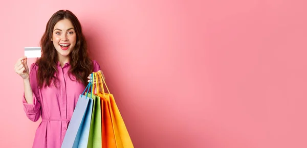 興奮した幸せなショッピングの肖像 ショッピングバッグを保持し プラスチッククレジットカードを示す女性は ピンクの背景に立って 驚いて笑って — ストック写真