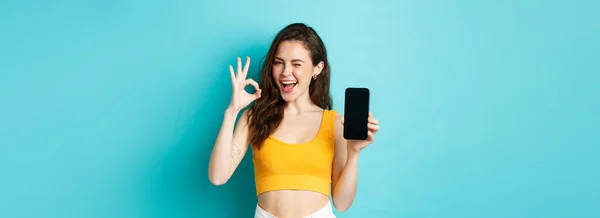 stock image Attractive modern woman in summer clothes, winking assuringly, showing okay sign with blank smartphone screen, showing your logo on display, blue background.