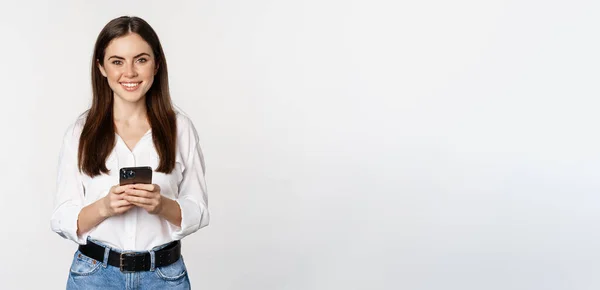 stock image Young woman with smartphone, smiling and looking at camera, using mobile phone app, cellular technology and online shopping concept, white background.