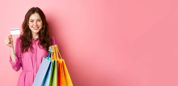 年轻的黑发女人拿着购物袋 拿着塑料信用卡笑着站在粉色的背景下 — 图库照片