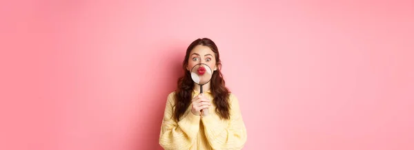 stock image Coquettish silly woman showing her pucker lips, kissing face with magnifying glass near mouth, staring funny at camera, standing against pink background.