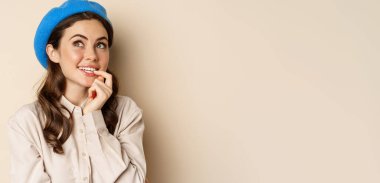 Close up portrait of dreamy smiling girl imaging, picturing smth, looking up and thinking, standing over beige background in blue trendy hat.