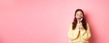 Gülümseyen genç bir kadın büyüteçle kusursuz beyaz dişli gülüşünü, diş sağlığı kliniğinin tanıtımını, stomatolojiyi ve sağlık hizmetlerini, pembe arka plana karşı duruşunu gösteriyor..