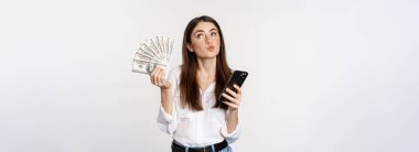 Portrait of happy smiling woman using mobile phone app, holding money cash, standing over white background. Copy space