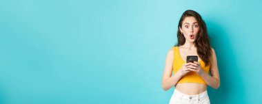 İlginç görünüyor. İlgi çekici güzel kız elinde akıllı telefon, heyecanla bakıyor ve online reklamları okuduktan sonra mavi arka planda duruyor..