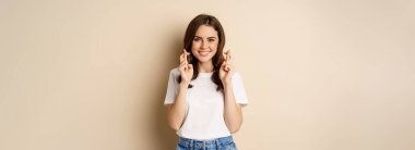 Hopeful woman praying, cross fingers for good luck, making wish, standing over beige background.