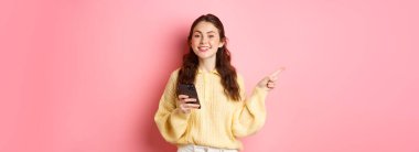 Güzel, modern bir kız akıllı telefonu elinde tutuyor ve bir kenara işaret ediyor. Cep telefonu kullanırken tanıtım metnini gösteriyor. Pembe arka planda duruyor..