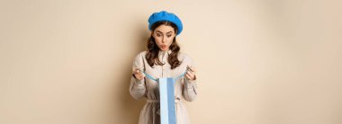 Image of girl looking surprised after opening gift bag, looking at present amazed, standing in stylish outfit over beige background.