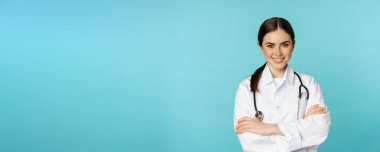 Medical staff and doctors concept. Young smiling female doctor, healthcare worker in white coat and stethoscope, looking confident, waiting patients, blue background.