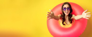 Close up portrait of beautiful happy woman inside pink swimming ring, luring, inviting people on summer vacation, beach party, standing over yellow background.