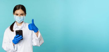Online doctor and clinic. Young woman in medical face mask, using smartphone for client remote online appointment, showing thumbs up, standing over blue background.