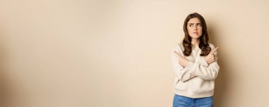 Complicated and indecisive woman pointing sideways, cant make choice, struggle with decision, choosing between two options, standing over beige background.