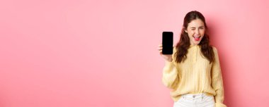 Küstah çekici bir kız görüntüsü, size akıllı telefon ekranını gösteriyor, göz kırpıyor ve gülümsüyor, cep telefonu öneriyor, pembe arka planda duruyor..