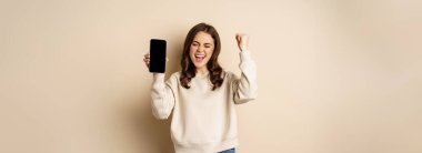 Happy woman winning, showing smartphone screen and shouting of joy, fist pump, standing over beige background.