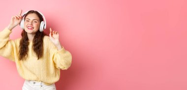 Güzel stil sahibi kadın model en sevdiği şarkıyı söylüyor, kablosuz kulaklıkla müzik dinliyor, gülümsüyor ve dans ediyor, pembe arka planda duruyor..