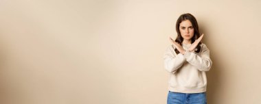 Woman showing stop, cross sign gesture and frowning angry, dislike and disapprove, saying no, rejecting and prohibiting, standing over beige background.