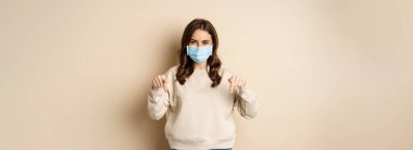 Cheerful cute woman in medical face mask, pointing fingers down, showing advertisement, using protection from covid-19, beige background.