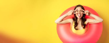 Portrait of attractive modern girl inside trendy pink beach swimming ring, showing peace v-sign and smiling happy, standing over yellow background.