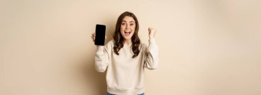 Enthusiastic woman cheering and showing cellphone screen, smartphone app interface, standing over beige background.